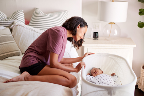 Mother kneeling on a bed looking at baby sleeping in a cot