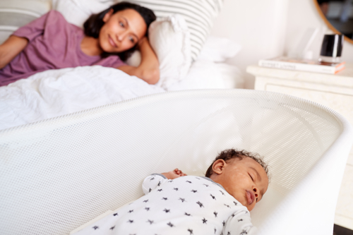 Mother lying on a bed looking at baby asleep in a cot